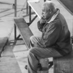 Elderly man sitting on bleachers, smoking and reflecting, captured in black and white.